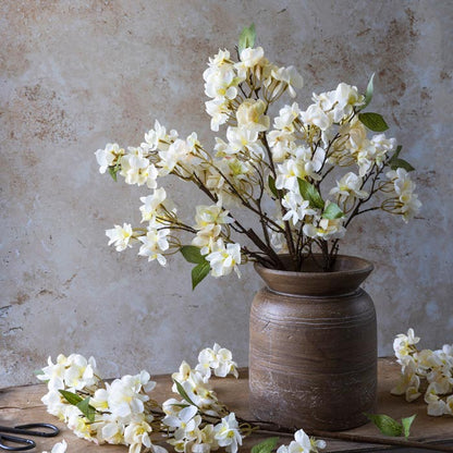A ceramic vase with a beige and brown color palette, featuring a design that mimics the natural curves of wood, filled with artificial white flowers and green foliage, set against a textured background.