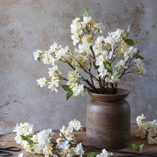A ceramic vase with a beige and brown color palette, featuring a design that mimics the natural curves of wood, filled with artificial white flowers and green foliage, set against a textured background.