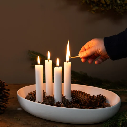 A hand is shown lighting a candle in a white matte candle holder, which is placed on a wooden surface. The holder contains four candles, and there is a bowl of decorative pine needles nearby.