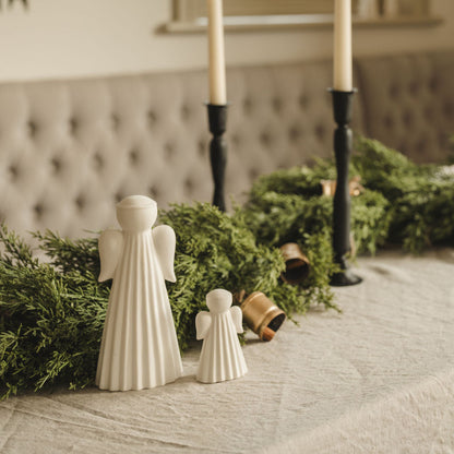 Two white angel figurines with a small child on a table with greenery and candles in the background.