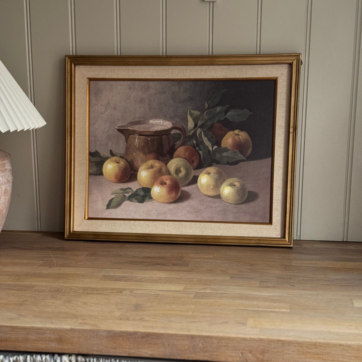 Framed still life painting of apples and a pitcher on a wooden surface with a lamp and striped fabric in the background.