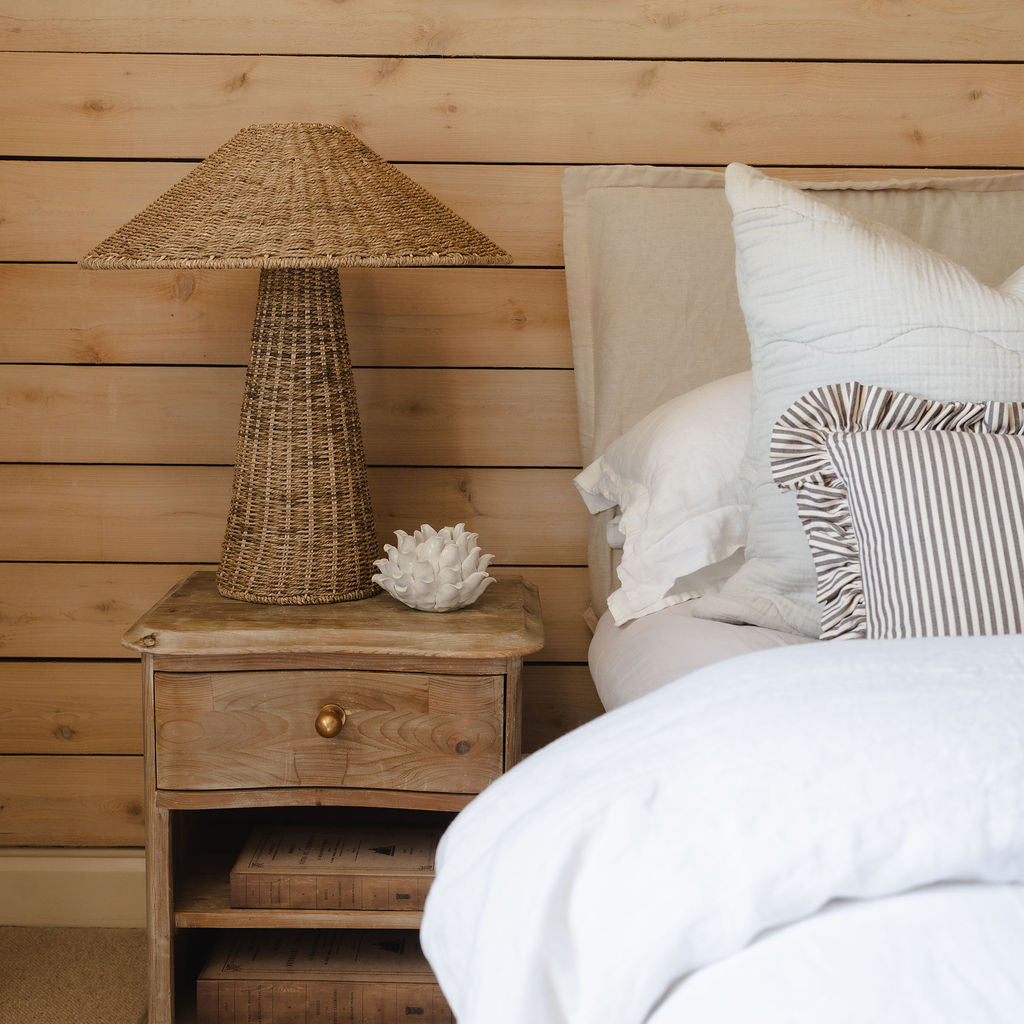 Wooden bedside table with lamp and decor next to a bed with white bedding and pillows.