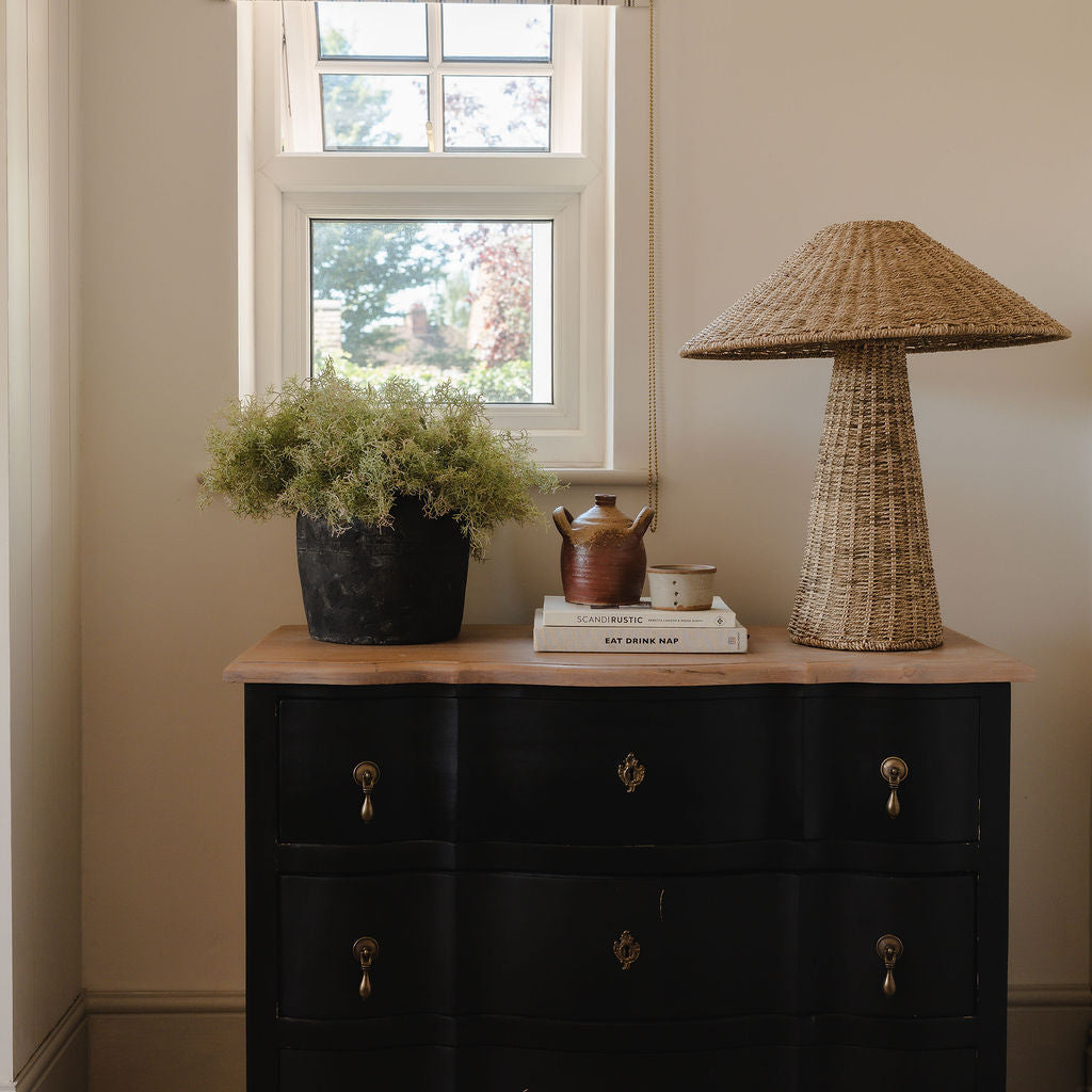 Black dresser with decorative items including a wicker lamp and potted plant, under a window with striped curtains.