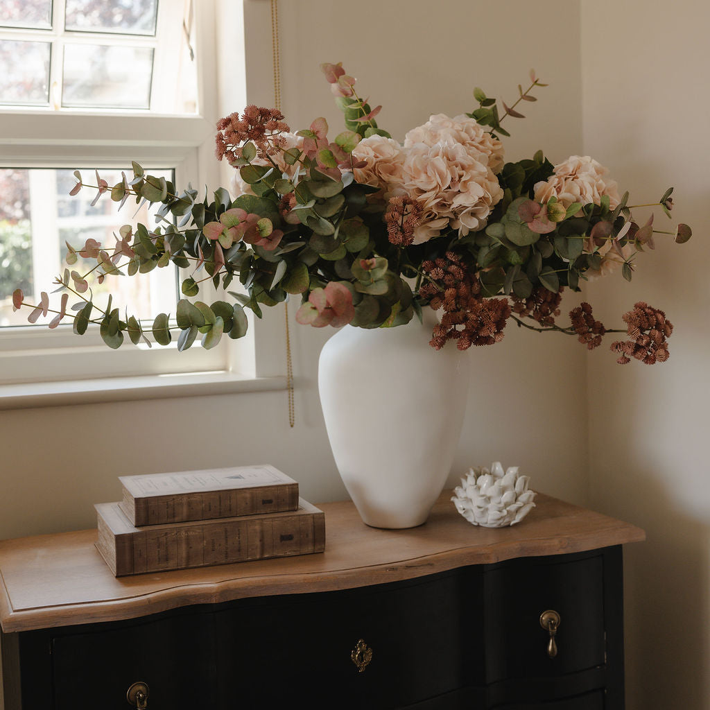Floral arrangement in a white vase on a wooden dresser with a window in the background.