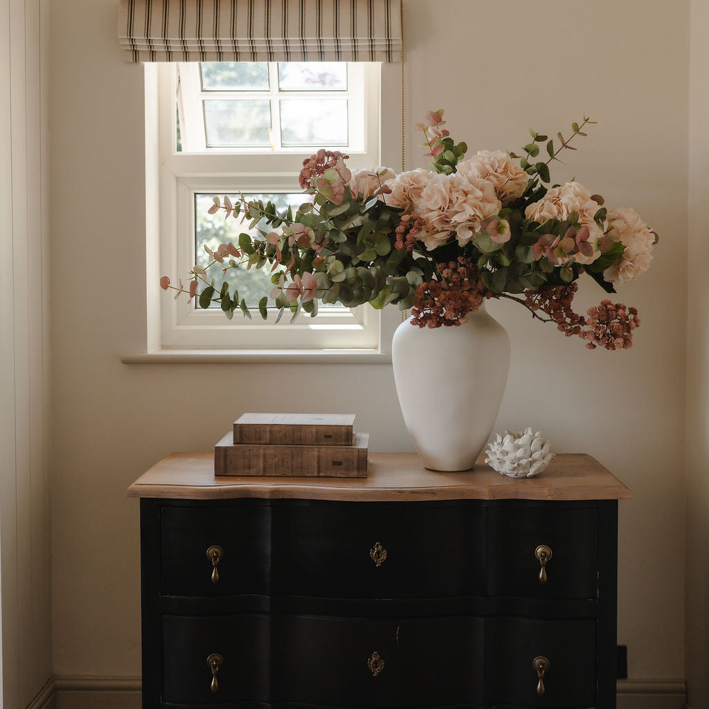 Decorative vase with flowers on a black dresser in a room with a window and light-colored walls.