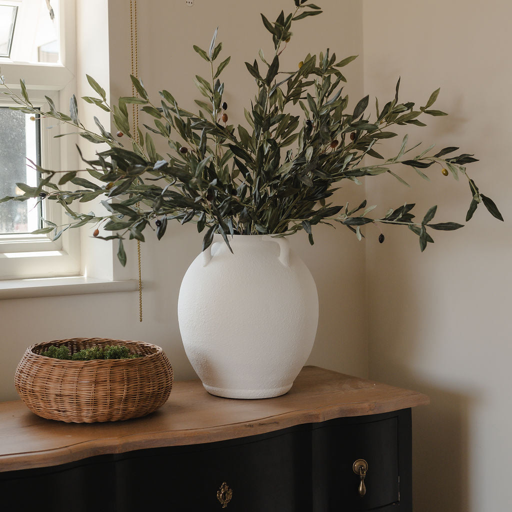 White vase with greenery on a wooden surface next to a window