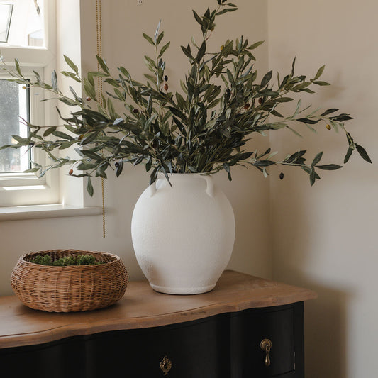 White vase with greenery on a wooden surface next to a window