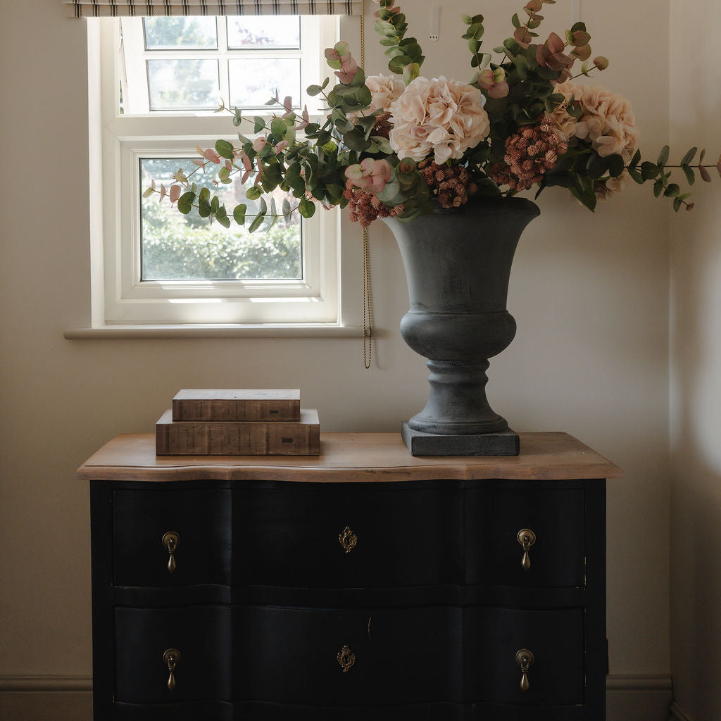 Floral arrangement in a gray urn on a black dresser by a window with a checkered curtain.
