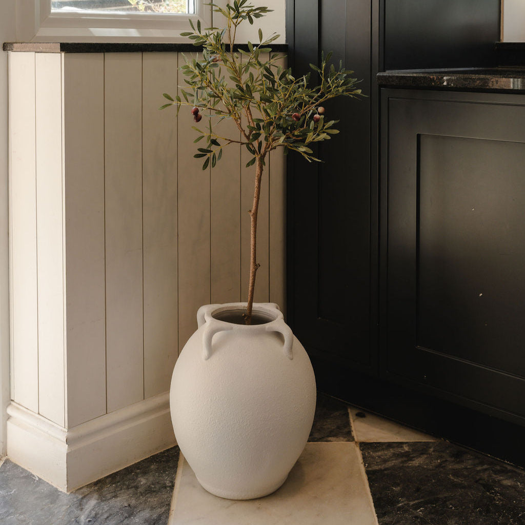White vase with a plant on a stone surface next to a window and dark cabinets.
