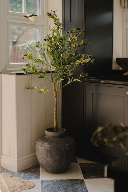 Faux olive tree with realistic green foliage in a weathered dark stone-effect planter, styled beside a window in a modern neutral-toned kitchen interior