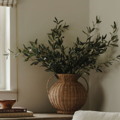White armchair with a patterned pillow, wicker vase with greenery, and wooden cabinet in a room.