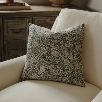 Decorative pillow with intricate pattern on a beige sofa, with a wooden side table and vase in the background.