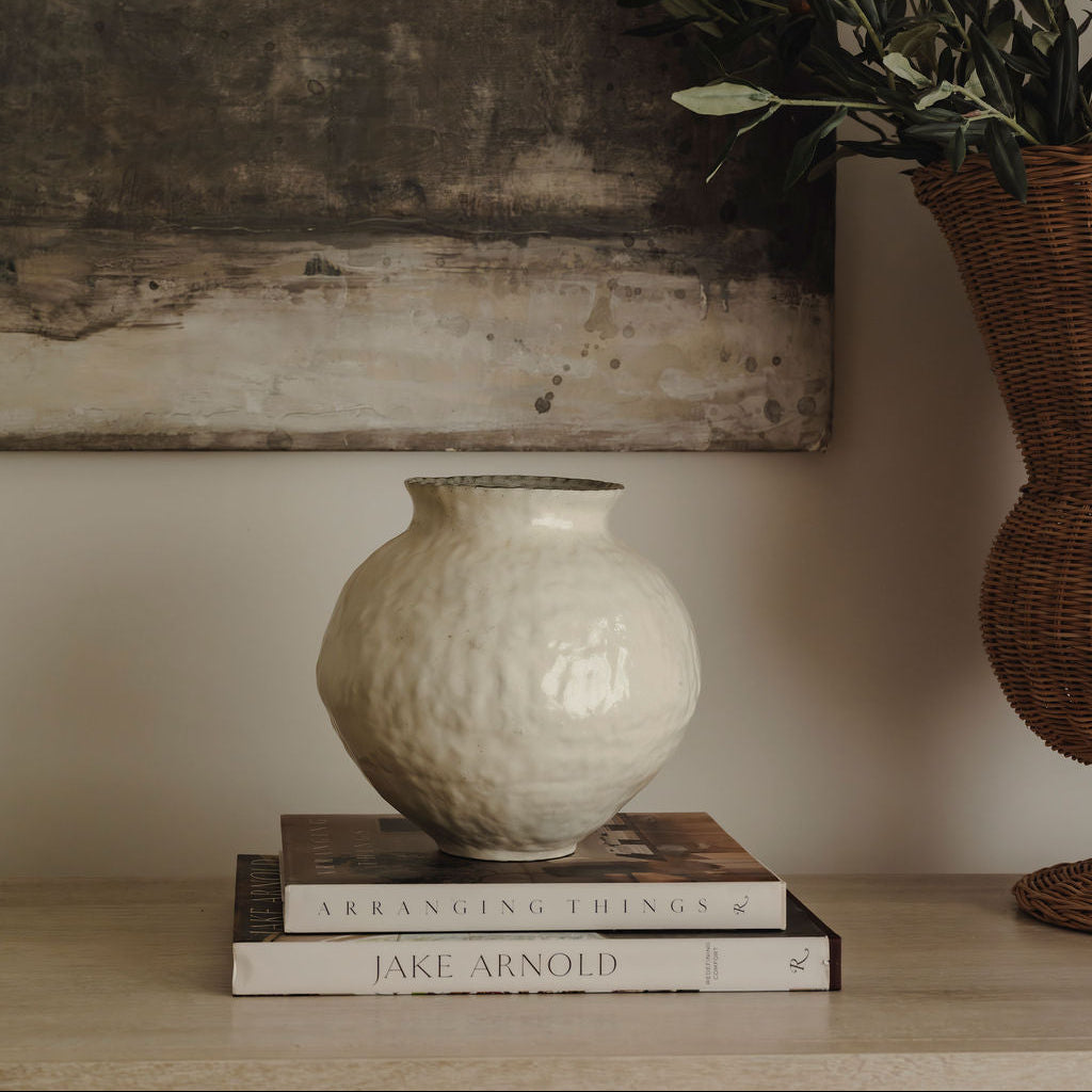 White textured vase on books with a wicker basket and plant against a textured wall.