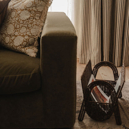 Brown sofa with a patterned pillow and a wicker magazine holder on a wooden floor.