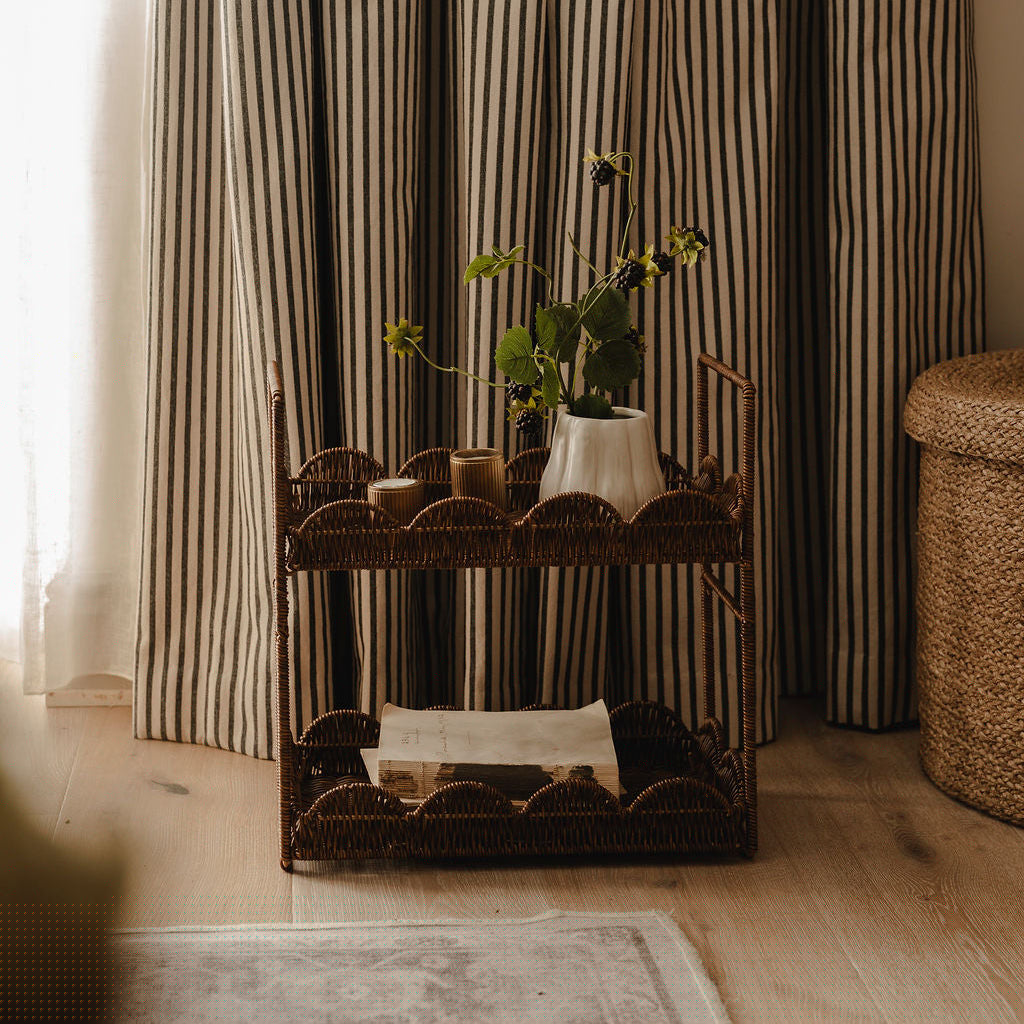 A rustic wicker two-tier cart styled in a cozy room with striped curtains, holding a white vase with green stems and small candles.