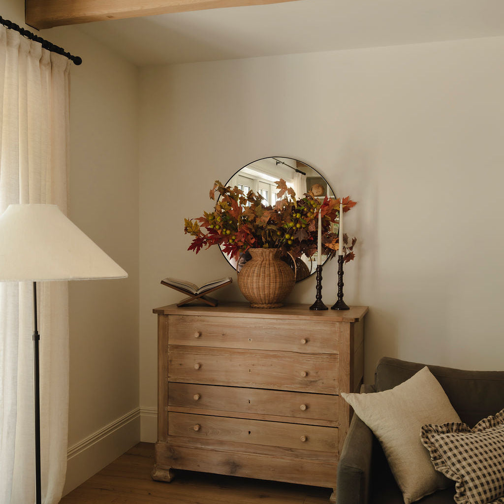 A rustic wooden three-drawer dresser in a warm beige finish, styled in a cozy living room setting with a round mirror, autumn flowers, a floor lamp, and a gray sofa.