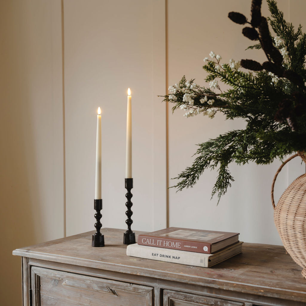 Two lit candles in black candlesticks on a wooden surface with books and a vase of flowers.