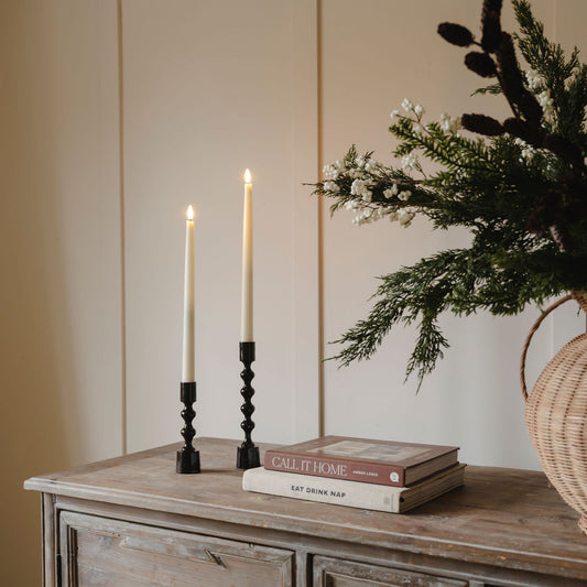 Two lit candles in black candlesticks on a wooden surface with books and a vase of flowers.